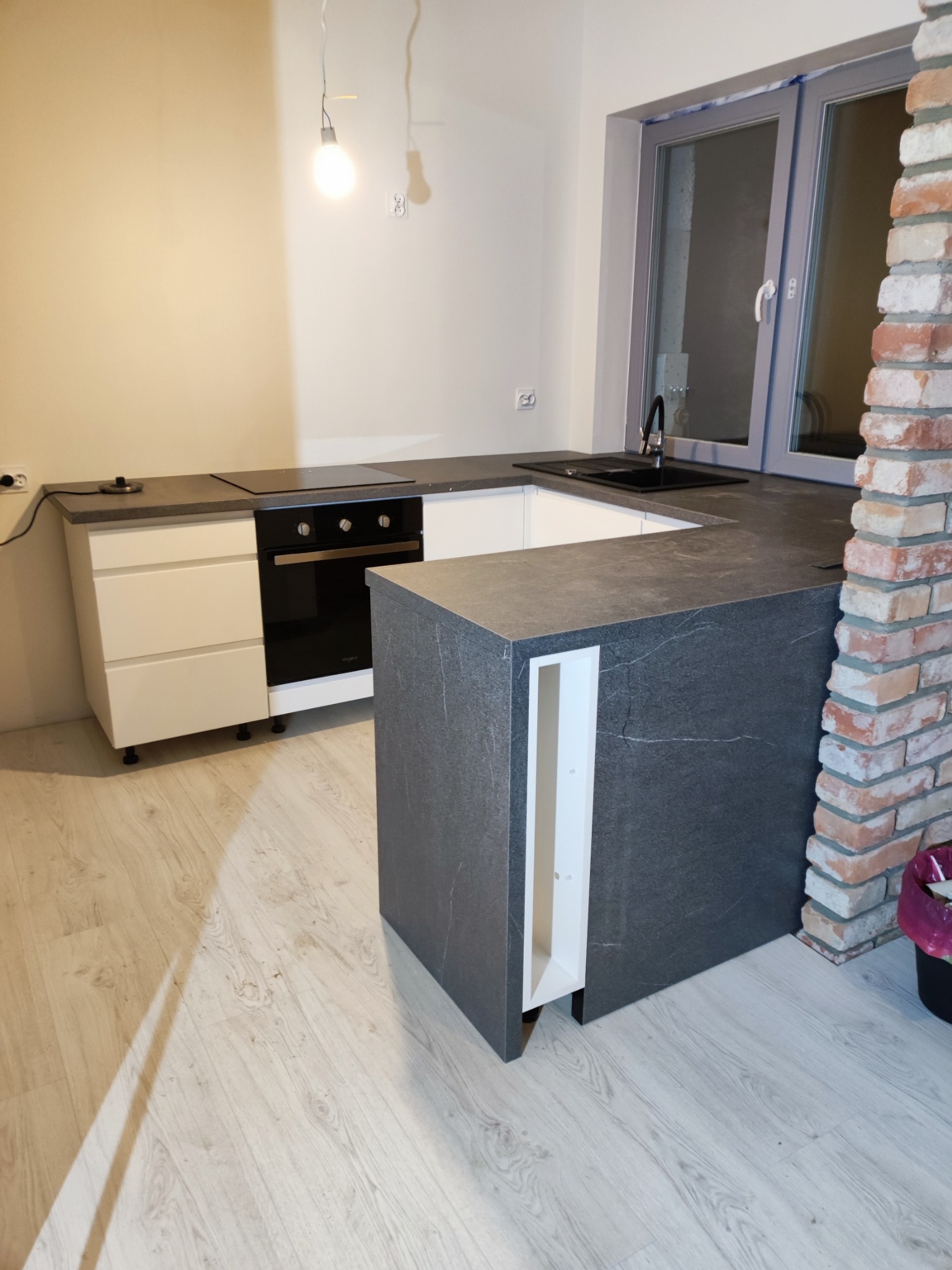 Montaż kuchni: wyspa kuchenna z szarym blatem imitującym kamień, białe szafki z piekarnikiem, okno z ceglanym obramowaniem, surowa ściana w tle, widoczna lampa wisząca.