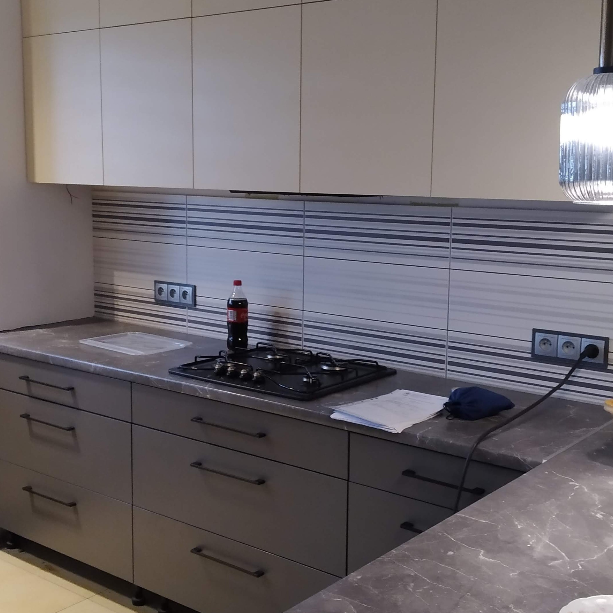 Szare szafki kuchenne z blatem imitującym marmur, płyta gazowa, butelka Coca-Coli i dokumenty na blacie, białe szafki wiszące, oświetlenie halogenowe, ściana wyłożona płytkami w poziome pasy.