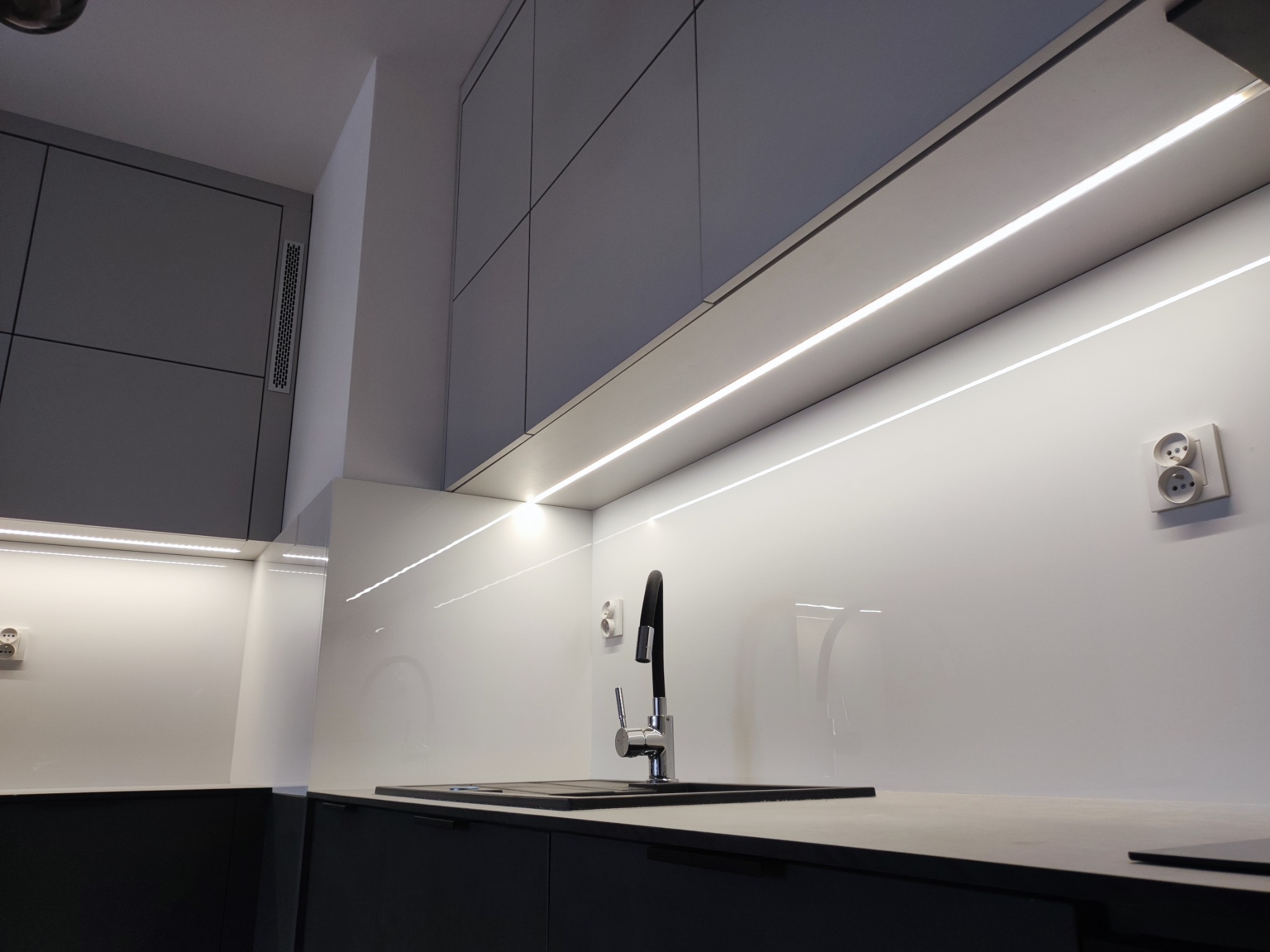 Nowoczesna kuchnia z szarymi szafkami, białym panelem szklanym nad blatem, oświetleniem LED podszafkowym i czarnym zlewem z baterią.