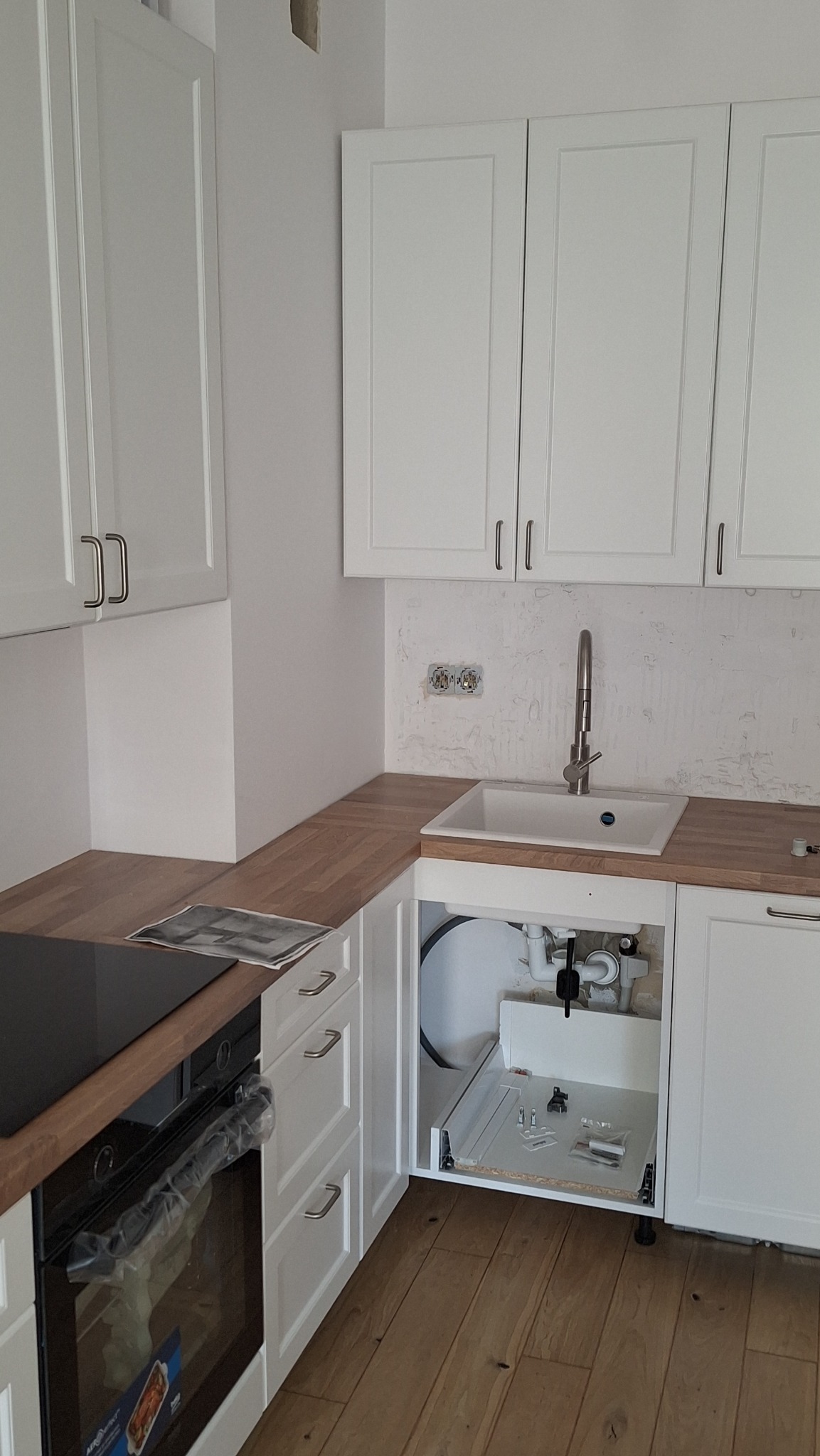 Montaż białej zabudowy kuchennej z drewnianym blatem, widoczne podłączenie zlewu i elementy instalacji w szafce, płyta indukcyjna z piekarnikiem obok.