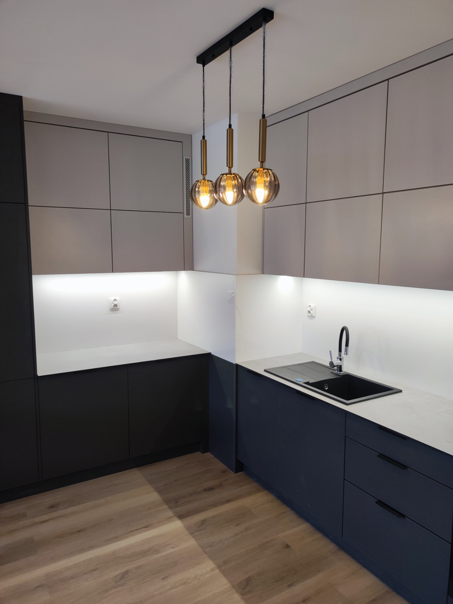 Nowoczesna kuchnia z szarymi górnymi szafkami i granatowymi dolnymi, oświetlona trzema wiszącymi lampami z kloszami w kształcie kuli, z drewnianą podłogą i czarnym zlewem.