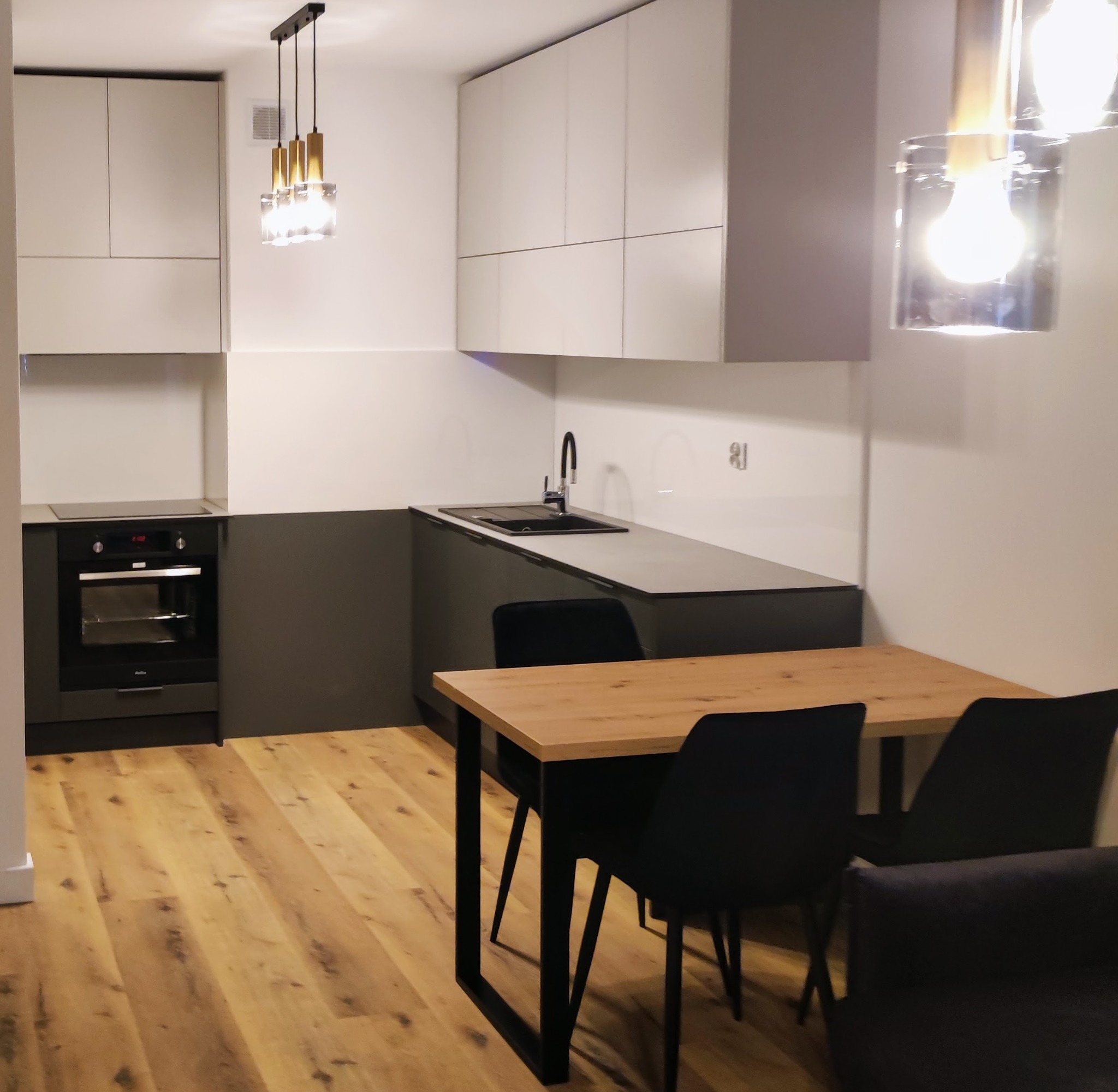 Nowoczesna kuchnia z szarymi szafkami dolnymi i białymi górnymi, drewnianym blatem i czarnymi krzesłami przy stole, oświetlona wiszącymi lampami z elementami złota.