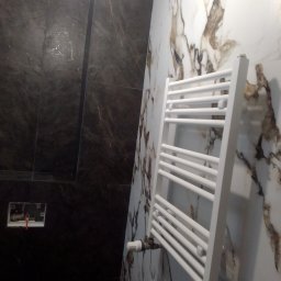 EWROMUR Andrii Gavryliuk - Biały grzejnik drabinkowy na tle marmurowych płytek w łazience. Widoczna instalacja i czarne płytki imitujące kamień. Ujęcie z dołu.