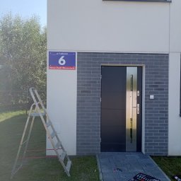 EWROMUR Andrii Gavryliuk - Nowoczesne drzwi wejściowe z szarym obramowaniem z cegły klinkierowej, skrzynka narzędziowa i drabina obok, adres budynku widoczny.