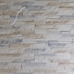 EWROMUR Andrii Gavryliuk - Ściana wyłożona dekoracyjną, beżowo-szarą okładziną imitującą kamień. Widoczny fragment białego kabla w lewym górnym rogu. Tekstura ściany z bliska.