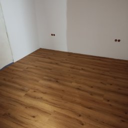 Ukończone układanie paneli podłogowych w rogu pomieszczenia z białymi ścianami. Widoczne gniazdka elektryczne bez osprzętu. Jasne, ciepłe odcienie drewna.