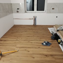 Ułożone panele podłogowe w jasnym odcieniu drewna w trakcie prac wykończeniowych. Widoczne rury instalacji, okno i fragment ściany z płytkami imitującymi marmur.