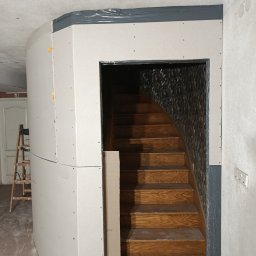 Montaż płyt gipsowo-kartonowych przy schodach z drewnianymi stopniami. Widoczna drabina i surowy stan wykończenia. Szare wykończenia ścian.