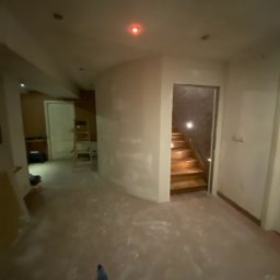 EWROMUR Andrii Gavryliuk - Wnętrze w trakcie prac wykończeniowych: ściany po szpachlowaniu, widoczne schody. Na podłodze resztki materiałów budowlanych i butelka. Oświetlenie punktowe.