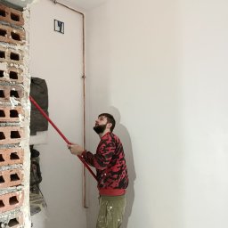 EWROMUR Andrii Gavryliuk - Mężczyzna maluje ścianę wałkiem na długim kiju w trakcie remontu. Widoczny fragment ściany z cegieł, instalacja miedziana i zakryty sprzęt budowlany.