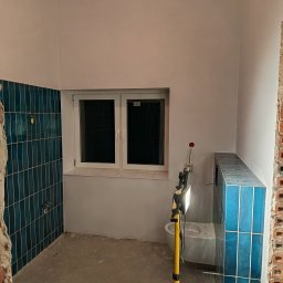 EWROMUR Andrii Gavryliuk - Remont łazienki w trakcie: ściany częściowo wyłożone pionowymi, turkusowymi płytkami, widoczne instalacje, okno i lampa budowlana. Surowy stan, brak fug.