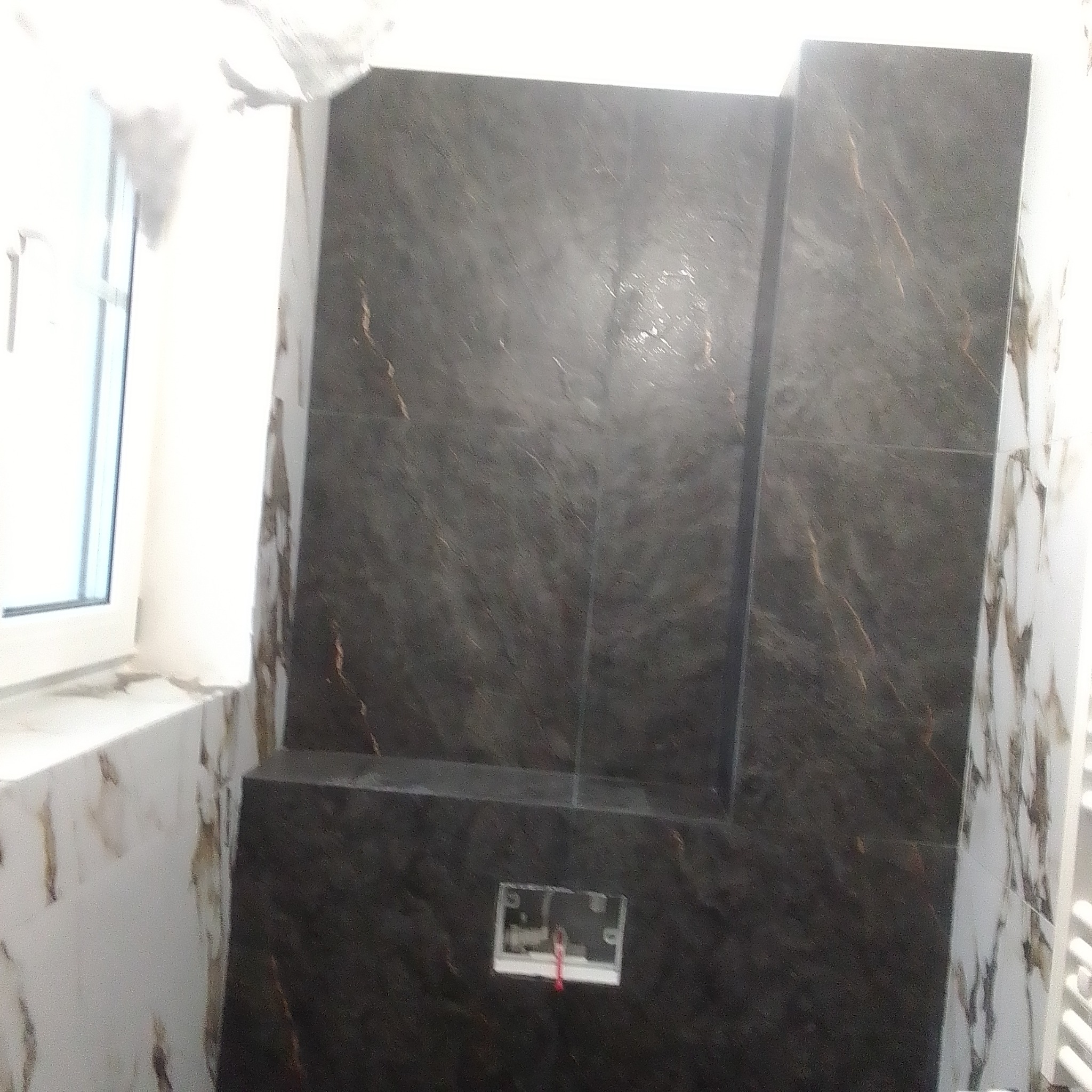 Ciemne, marmurowe płytki ścienne z złotymi żyłkami w łazience, częściowo ułożone. Widoczna instalacja podtynkowa. Białe płytki z marmurowym wzorem przy oknie.