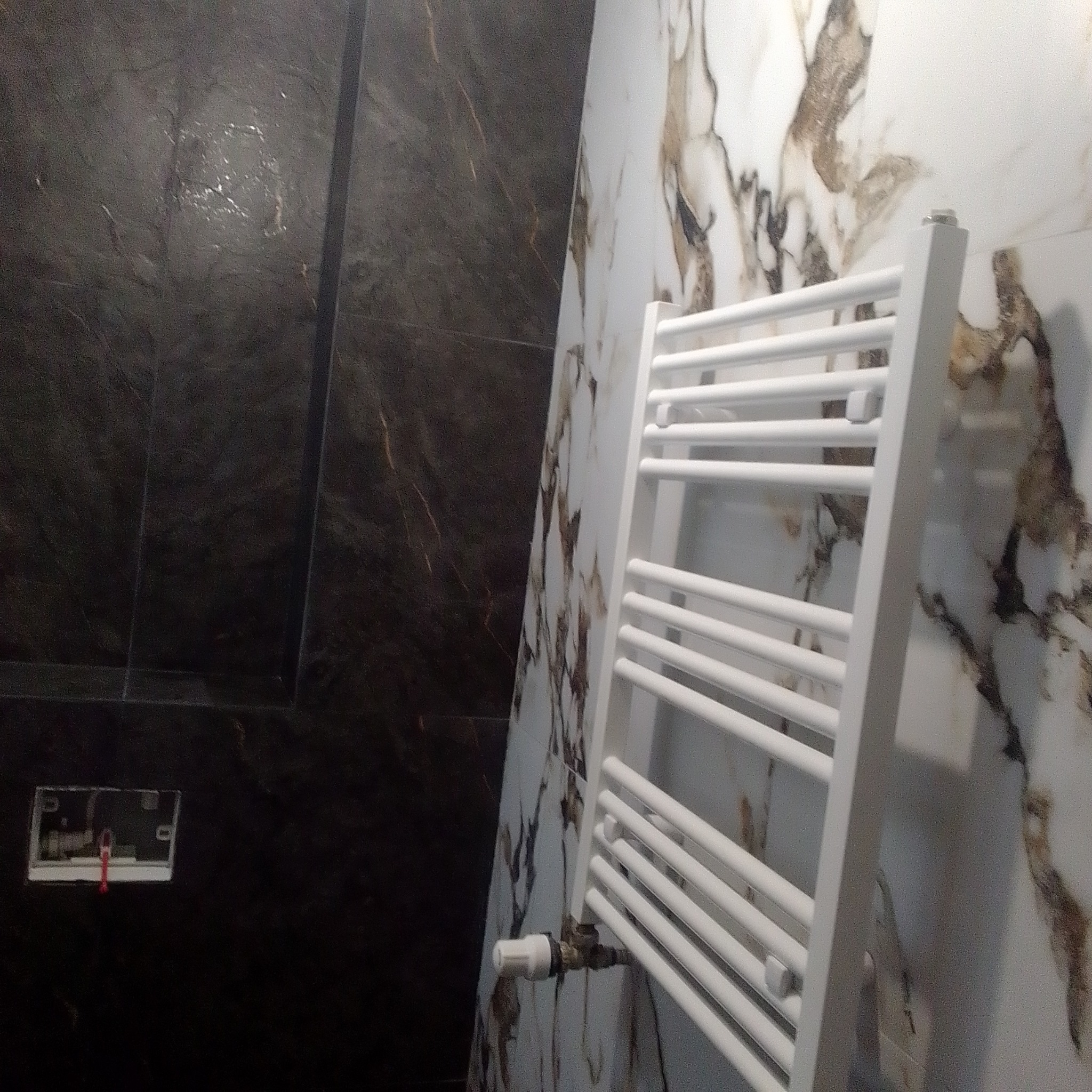 Biały grzejnik drabinkowy na tle marmurowych płytek w łazience. Widoczna instalacja i czarne płytki imitujące kamień. Ujęcie z dołu.