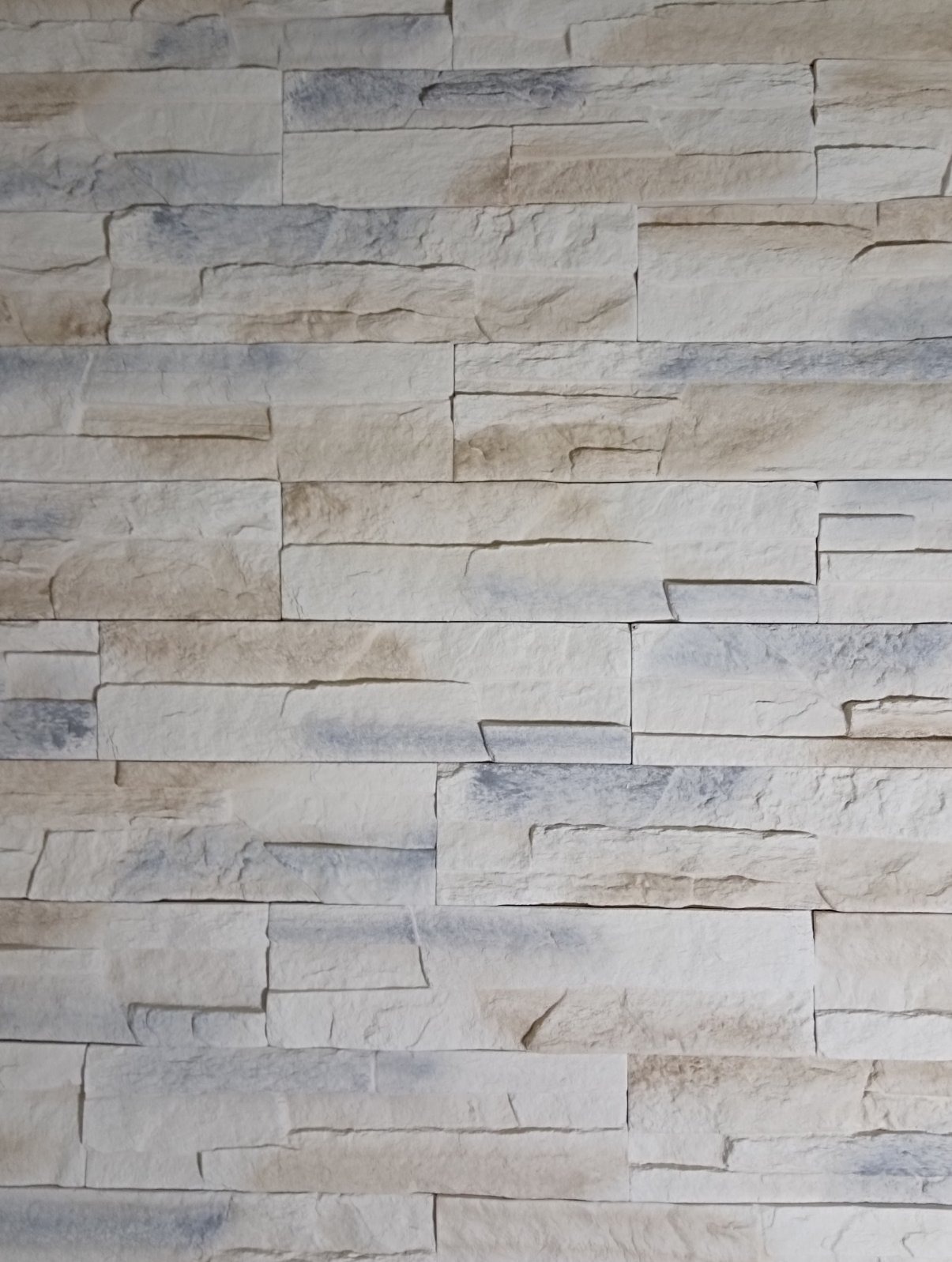 Ściana wykończona dekoracyjną, jasną okładziną imitującą kamień. Widoczne nieregularne kształty i subtelne odcienie beżu i szarości, tworzące trójwymiarowy efekt.