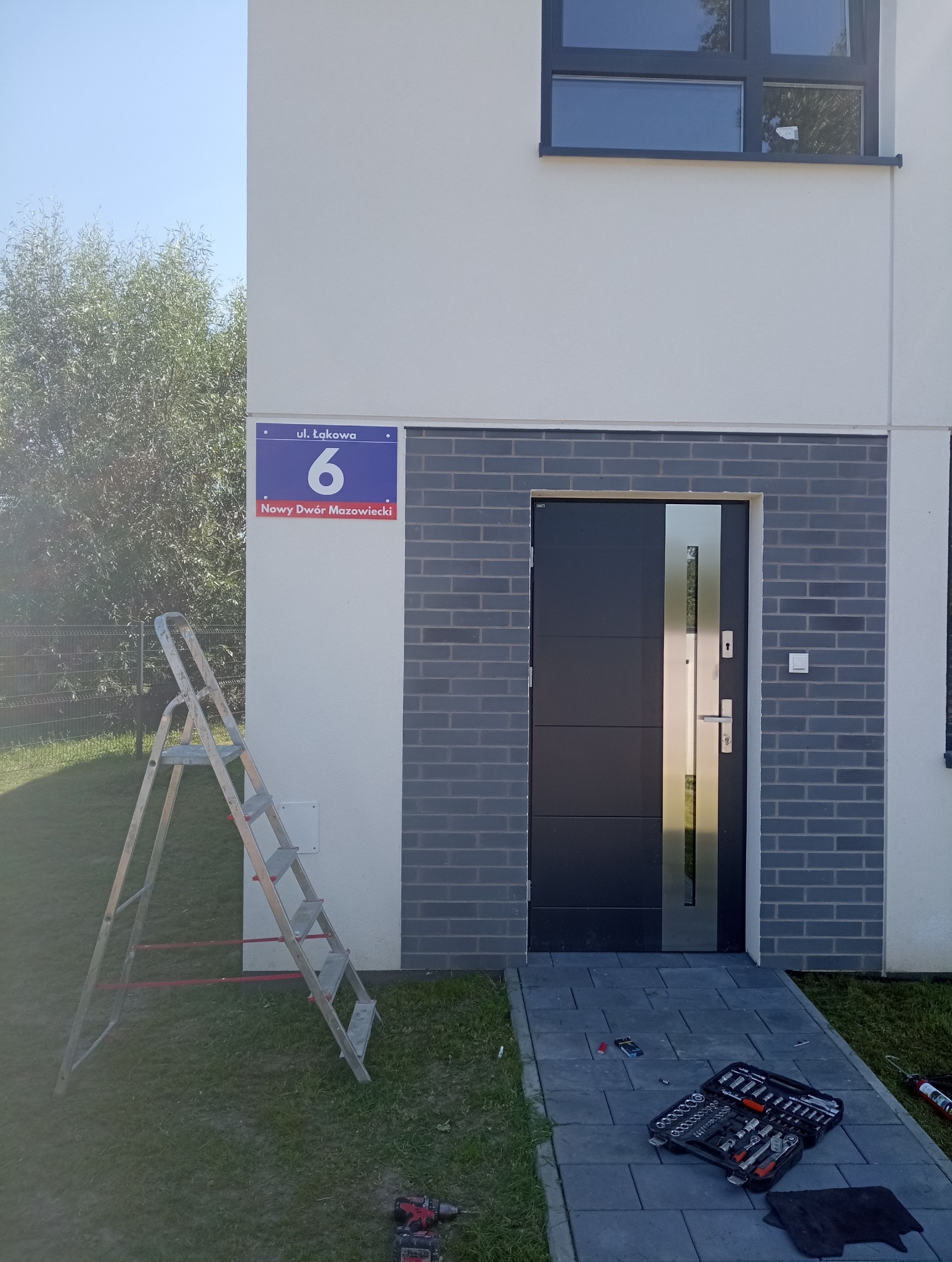 Nowoczesne drzwi wejściowe z szarym obramowaniem z cegły klinkierowej, skrzynka narzędziowa i drabina obok, adres budynku widoczny.