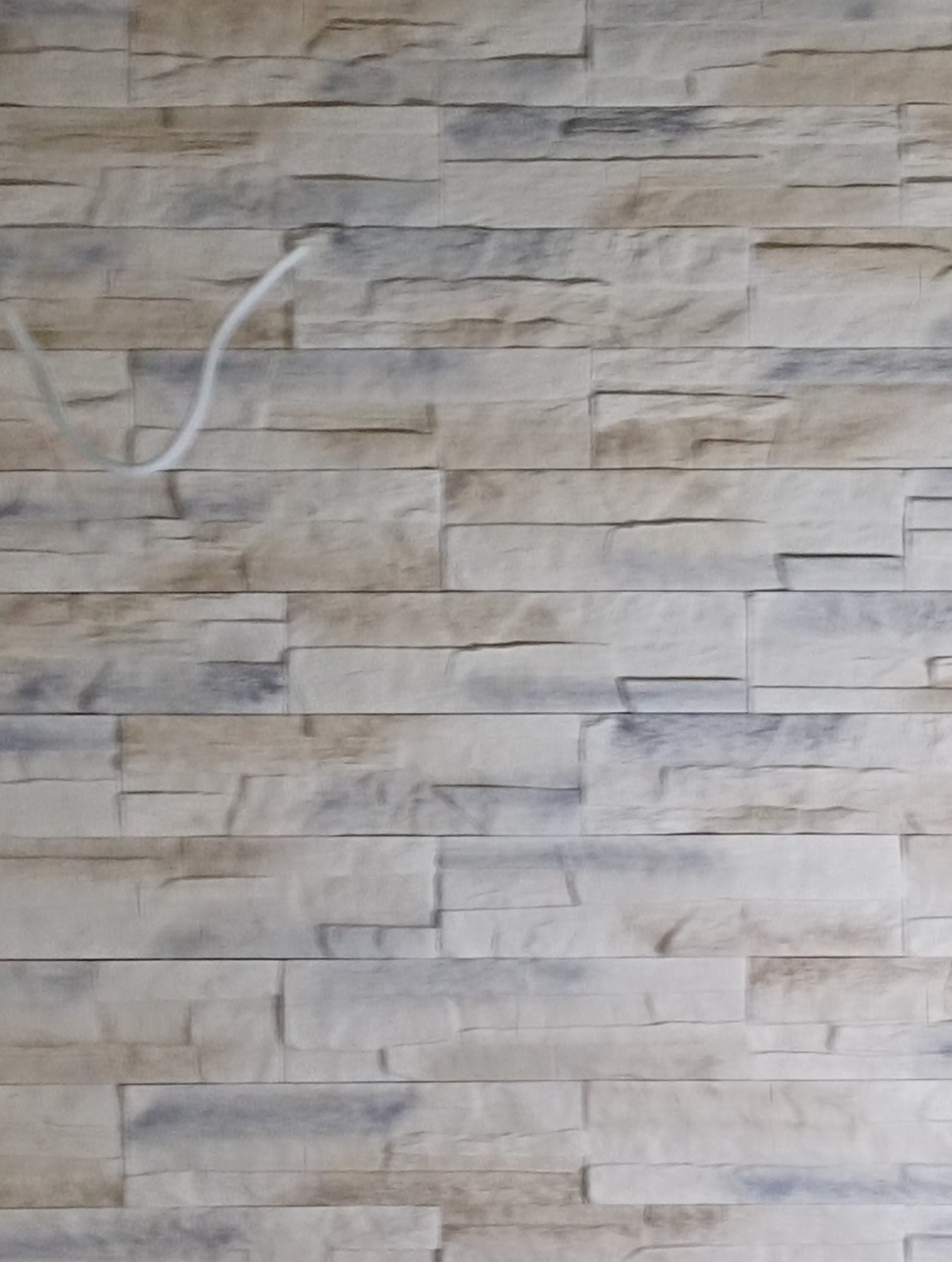 Ściana wyłożona dekoracyjną, beżowo-szarą okładziną imitującą kamień. Widoczny fragment białego kabla w lewym górnym rogu. Tekstura ściany z bliska.