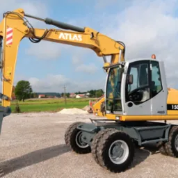 Żółto-szara maszyna budowlana Atlas 1505 M na tle pola i domów, widok z boku na żwirowym podłożu.