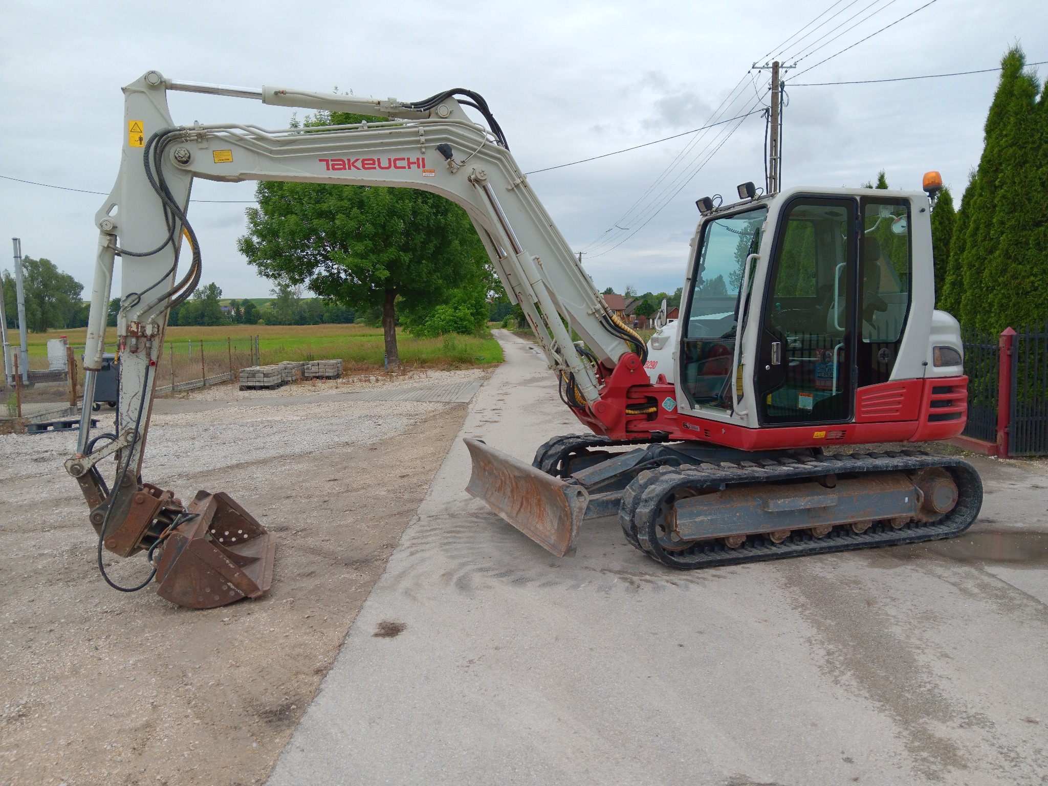 Minikoparka Takeuchi w Zagajowie, zaparkowana na drodze z opuszczoną łyżką i lemieszem, widoczne ślady użytkowania.
