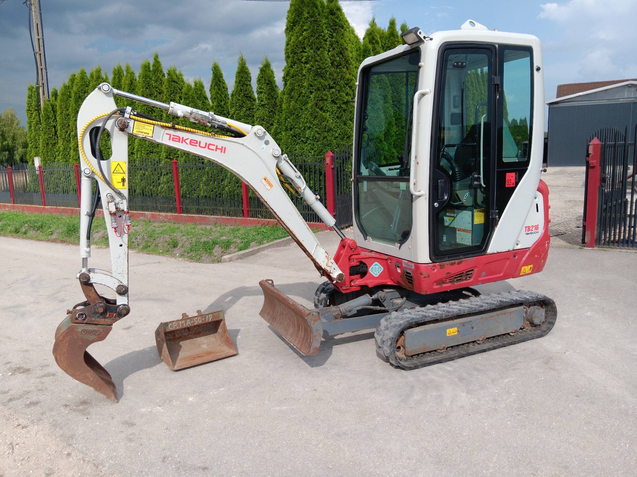 Mała, używana maszyna budowlana Takeuchi TB216 z łyżką i lemieszem, zaparkowana na betonowym placu, widoczne ślady użytkowania.