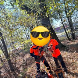 Green Forest Marcin Warchoł - Dwóch pracowników w pomarańczowych uniformach, z zasłoniętymi twarzami, trzyma piły spalinowe Husqvarna w lesie, obok stoją torby na narzędzia, w tle widać drzewa i budynek.