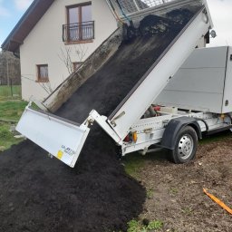 Green Forest Marcin Warchoł - Wyładunek czarnej ziemi z wywrotki na trawnik przed domem z balkonem, widoczna łopata wewnątrz skrzyni ładunkowej.