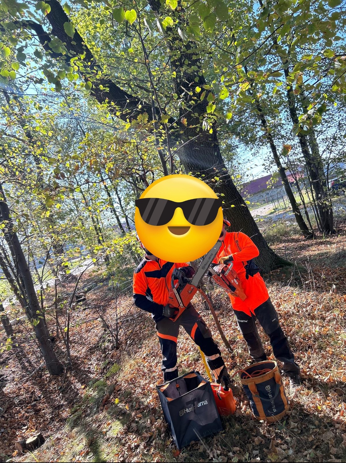 Dwóch pracowników w pomarańczowych uniformach, z zasłoniętymi twarzami, trzyma piły spalinowe Husqvarna w lesie, obok stoją torby na narzędzia, w tle widać drzewa i budynek.