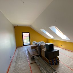 Na Zicher Group - Pokój na poddaszu po malowaniu: żółta ściana z oknami dachowymi, szara ściana boczna, podłoga zabezpieczona folią, materiały budowlane i rusztowanie w centrum.