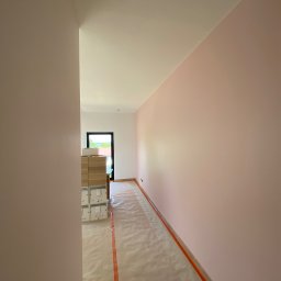 Na Zicher Group - Świeżo pomalowany korytarz w odcieniach różu, zabezpieczona podłoga folią i taśmą, kartony w tle, widok na okno z zielenią.