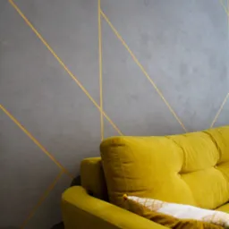 Szara ściana z geometrycznym wzorem w złotym kolorze, widoczny fragment żółtej sofy z poduszką. Nowoczesny design wnętrza.