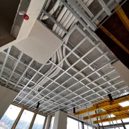 Na Zicher Group - Sufit z metalową konstrukcją, widoczne rury i kable, częściowo zamontowana płyta. Ujęcie od dołu, okna w tle. Widać elementy rusztowania.