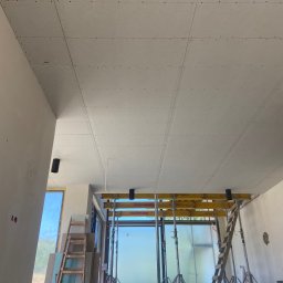 Na Zicher Group - Montaż płyt na suficie z widocznymi podporami i drabiną. Surowy stan wykończenia wnętrza, duże okno w tle.