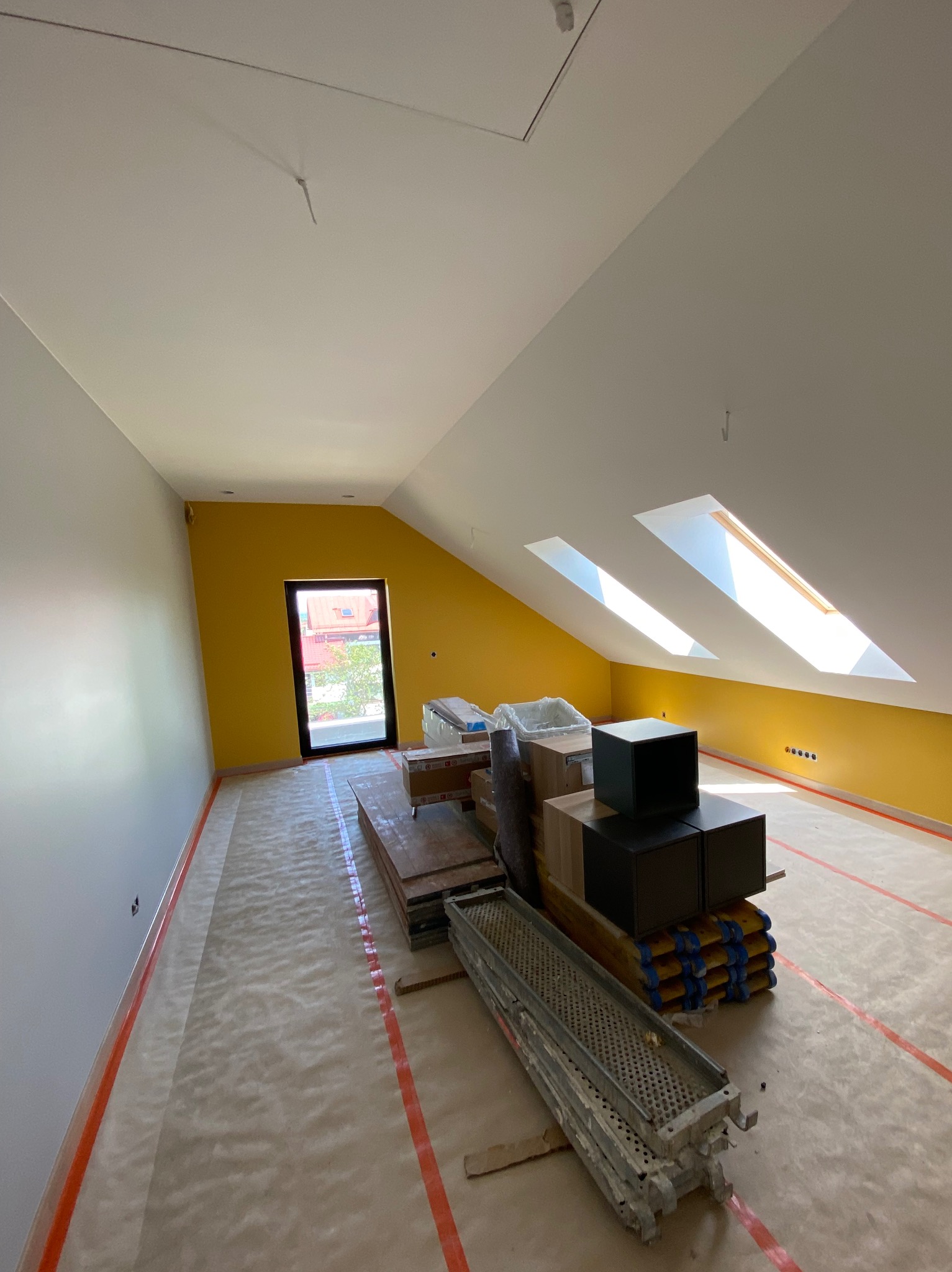 Pokój na poddaszu po malowaniu: żółta ściana z oknami dachowymi, szara ściana boczna, podłoga zabezpieczona folią, materiały budowlane i rusztowanie w centrum.