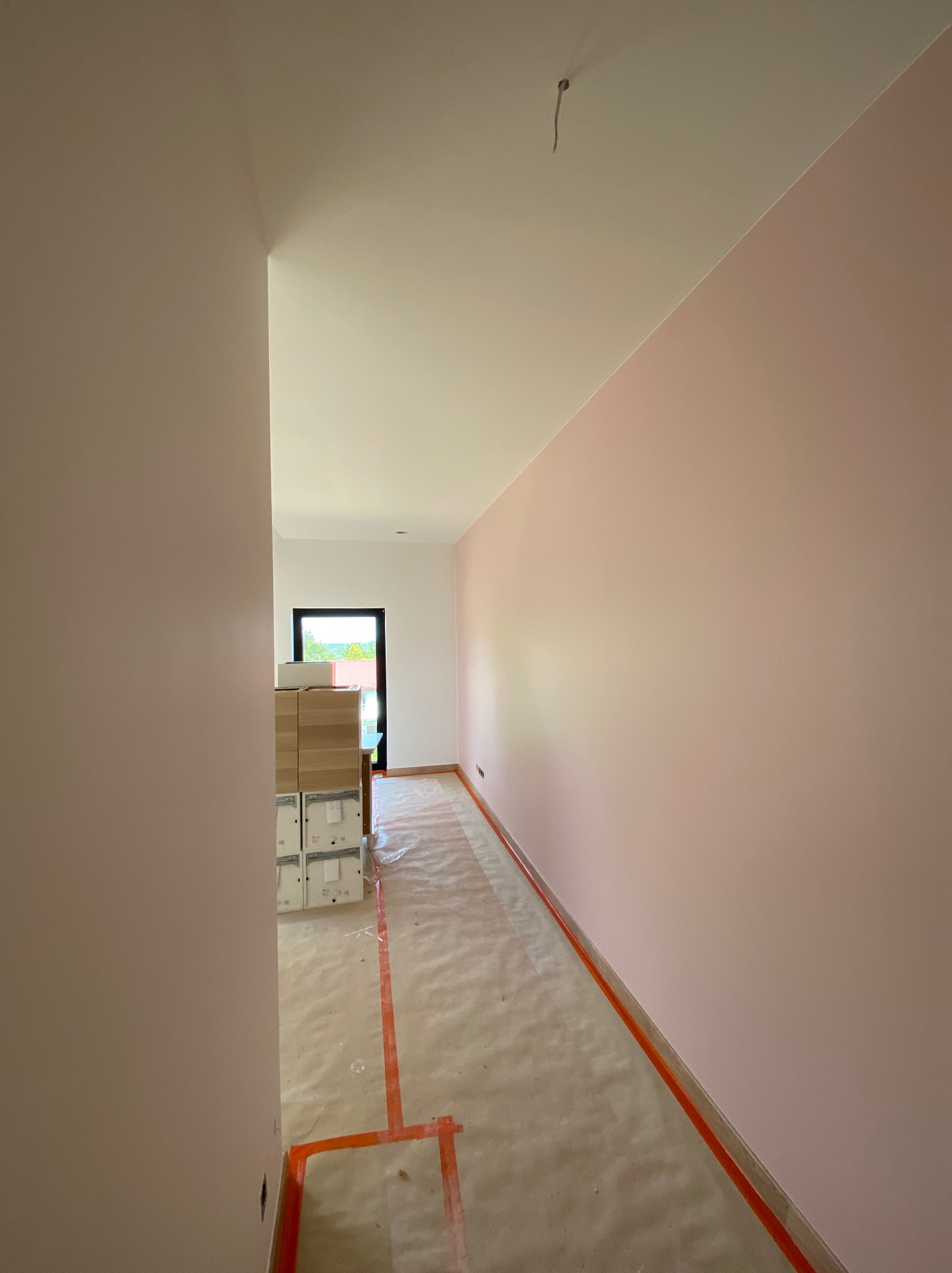 Świeżo pomalowany korytarz w odcieniach różu, zabezpieczona podłoga folią i taśmą, kartony w tle, widok na okno z zielenią.