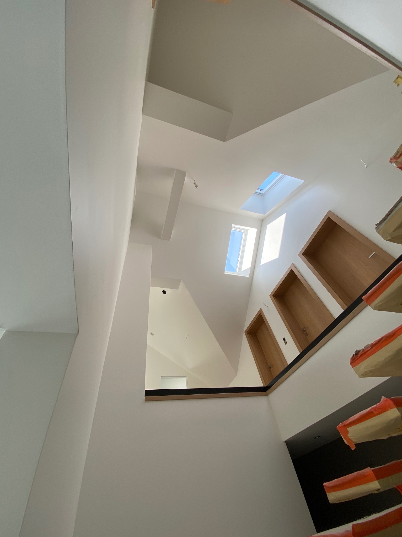 Wnętrze domu w trakcie prac. Widok na klatkę schodową z oknem dachowym i drewnianymi drzwiami. Schody zabezpieczone folią, ściany świeżo pomalowane na biało.