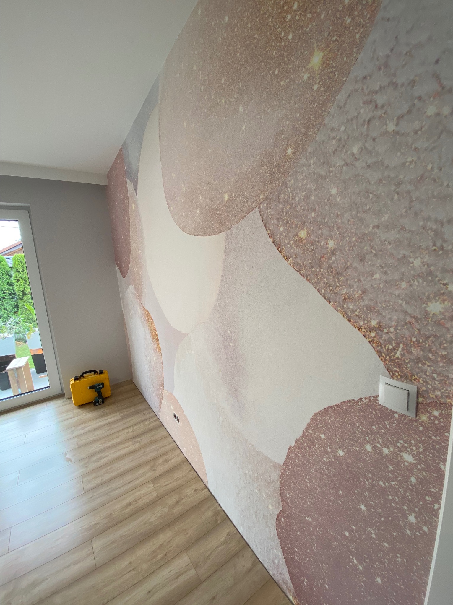 Ściana z tapetą w abstrakcyjne, różowe wzory, widoczny włącznik światła i skrzynka z narzędziami przy podłodze. Parkiet w jasnym odcieniu drewna.