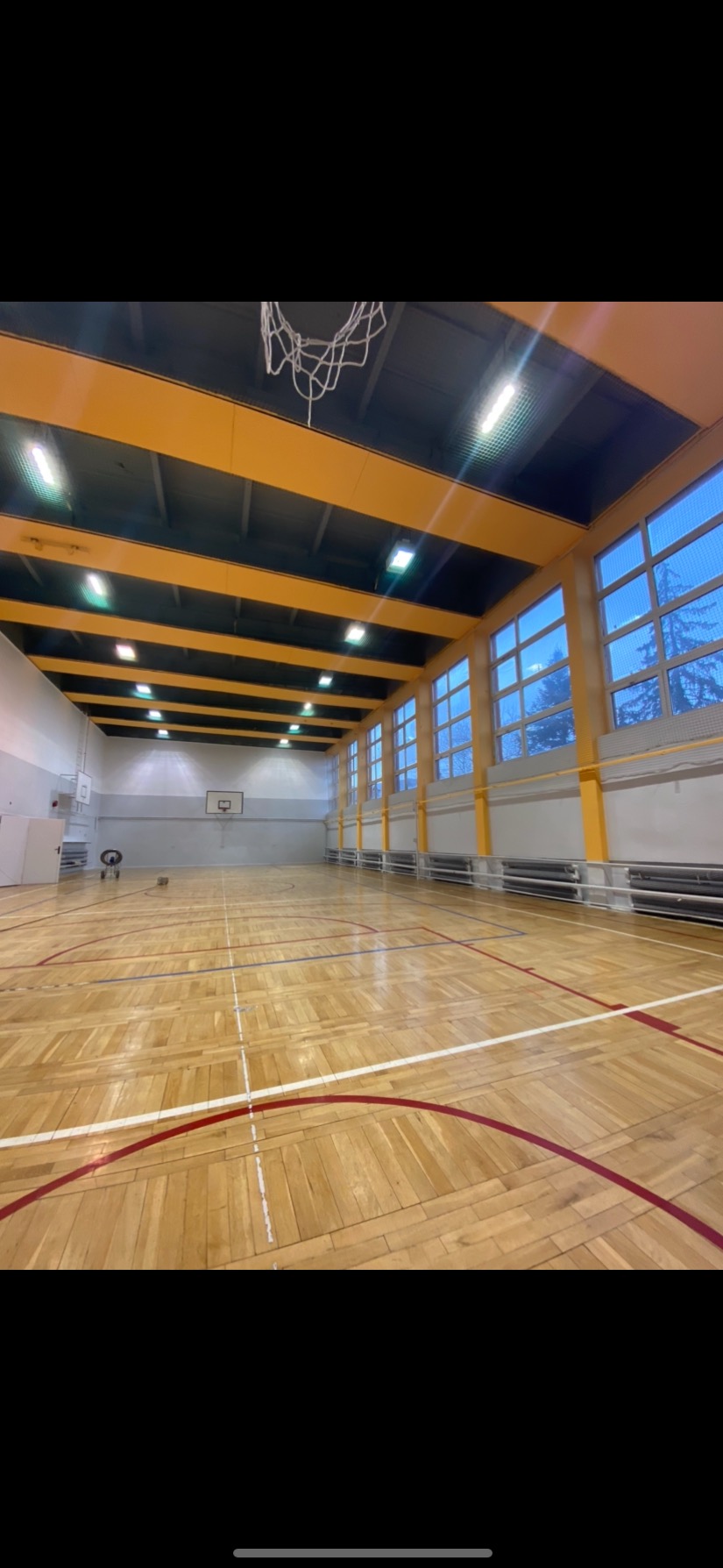 Wnętrze jasnej sali gimnastycznej z parkietem w geometryczne wzory, żółtymi belkami stropowymi i wysokimi oknami w tle, widok na kosz do koszykówki.