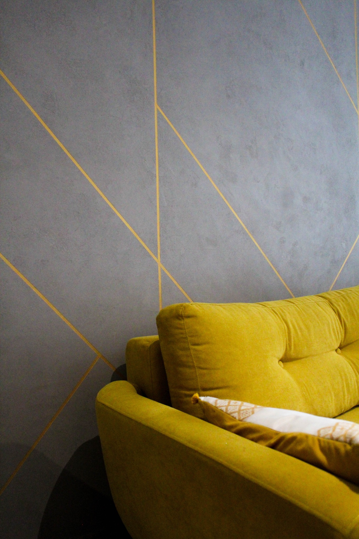 Szara ściana z geometrycznym wzorem w złotym kolorze, widoczny fragment żółtej sofy z poduszką. Nowoczesny design wnętrza.
