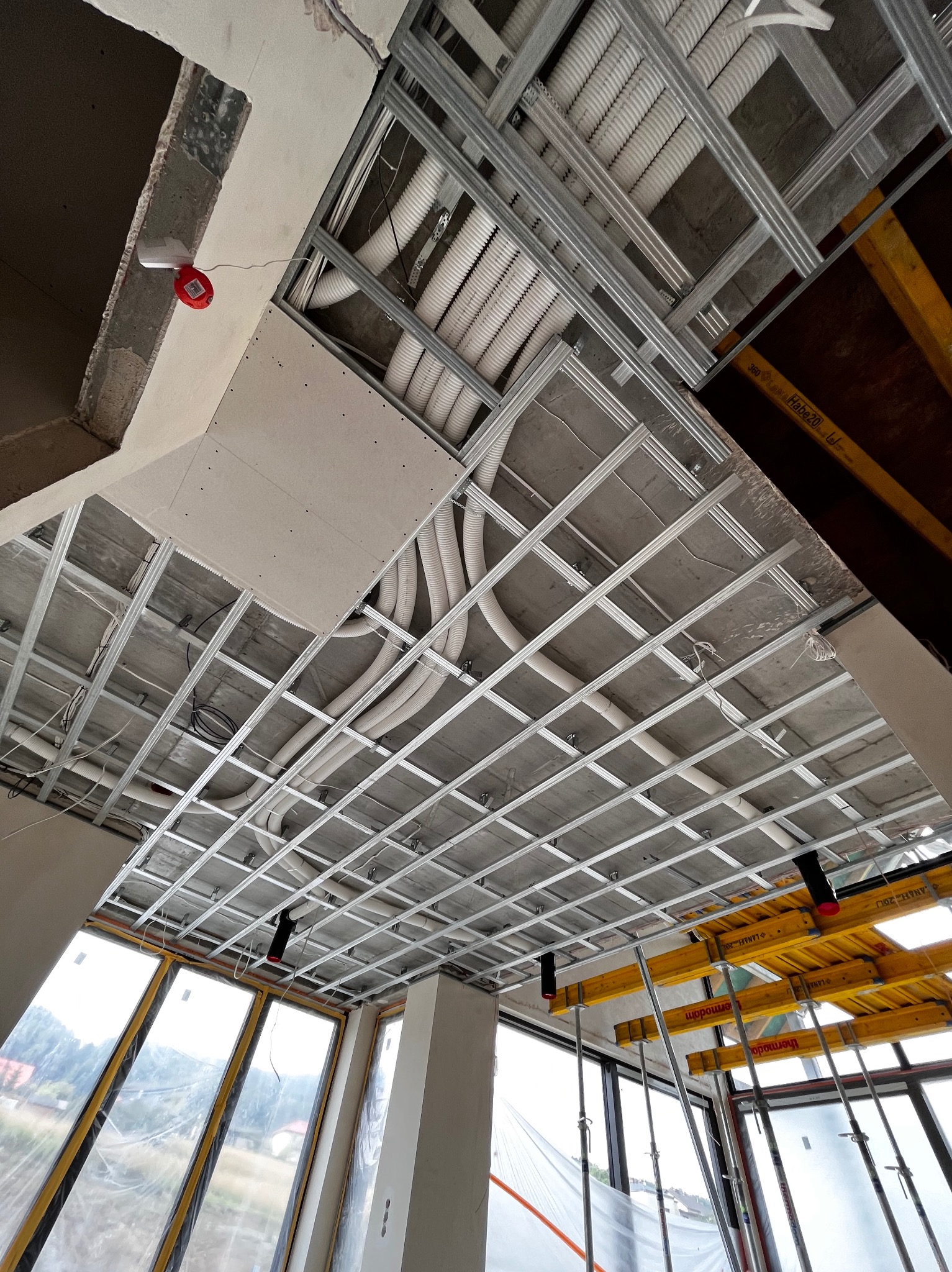 Sufit z metalową konstrukcją, widoczne rury i kable, częściowo zamontowana płyta. Ujęcie od dołu, okna w tle. Widać elementy rusztowania.