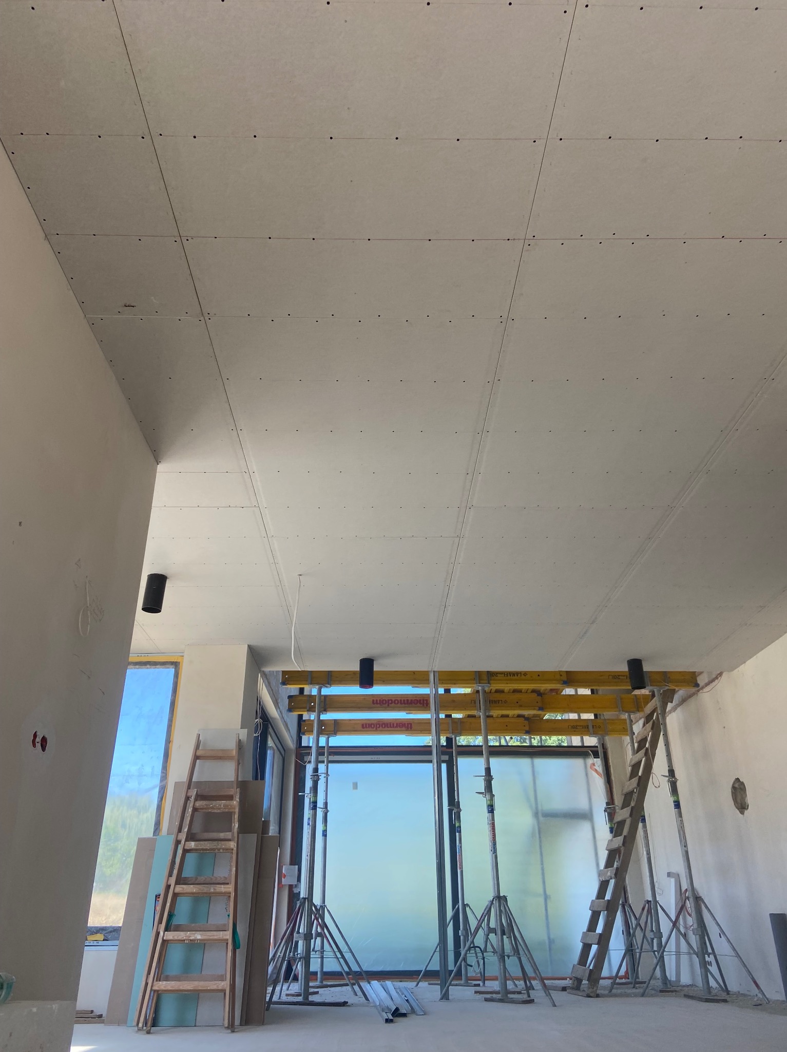 Montaż płyt na suficie z widocznymi podporami i drabiną. Surowy stan wykończenia wnętrza, duże okno w tle.
