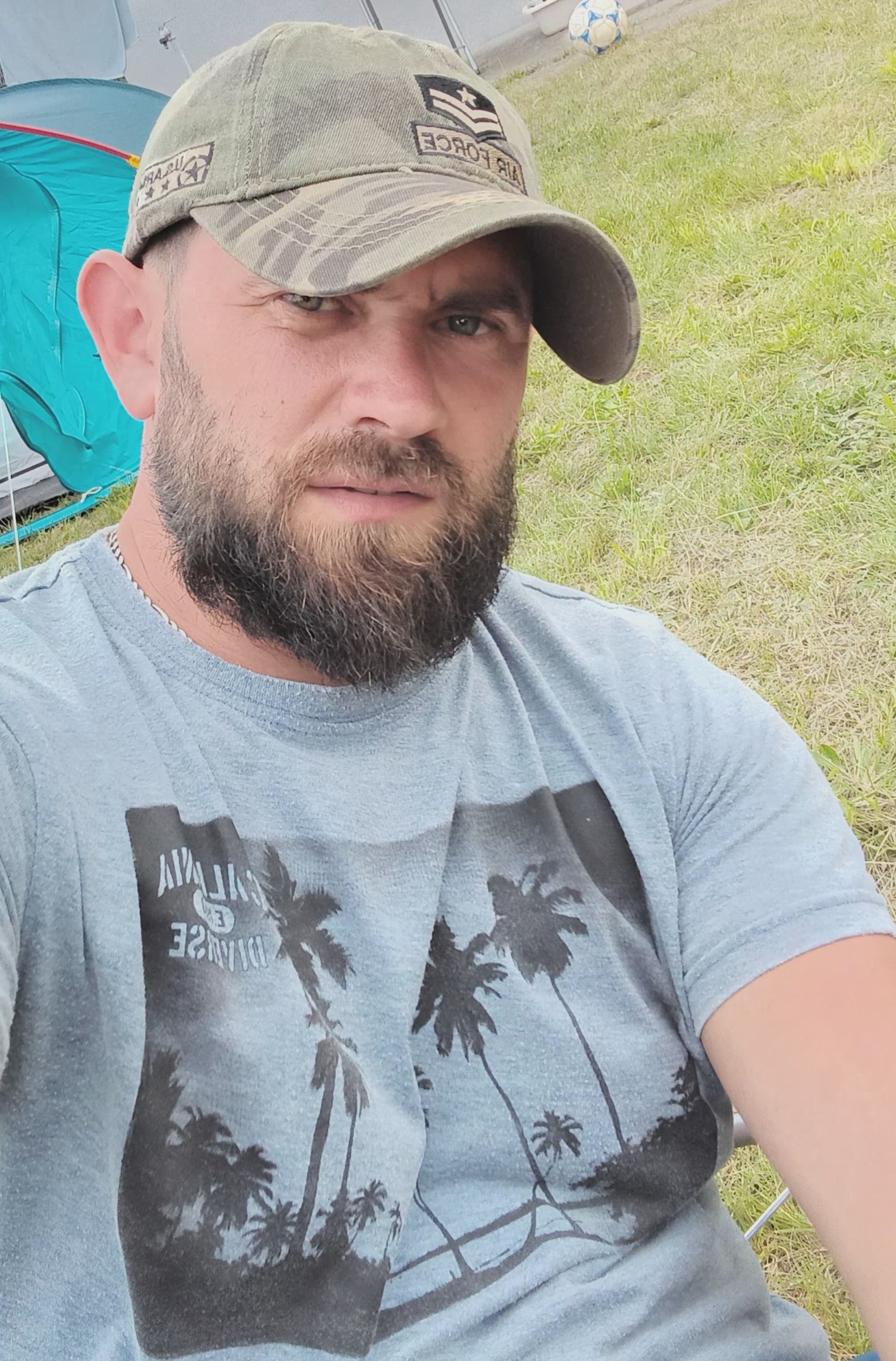 Mężczyzna z brodą w czapce z daszkiem, na tle trawy i namiotu, z piłką w tle.