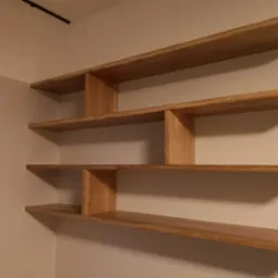 Asymetryczne, wiszące p&oacute;łki z jasnego drewna w minimalistycznym wnętrzu, mocowane do ściany, z widocznymi naturalnymi słojami drewna i subtelnym oświetleniem.