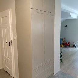 Woodland - Wysoka, szara szafa wnękowa z frezowanymi frontami, dopasowana kolorystycznie do ściany, z widocznymi trzema szufladami w dolnej części i białymi drzwiami po lewej stronie, w tle pokój dziecięcy...