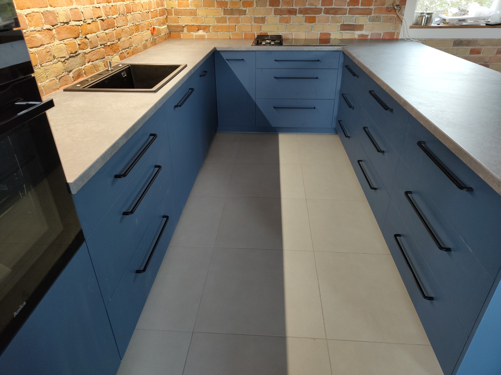 U-kształtny układ szafek kuchennych w odcieniach niebieskiego z czarnymi, podłużnymi uchwytami, szary blat i ceglana ściana w tle, nowoczesna płyta grzewcza i zlew.