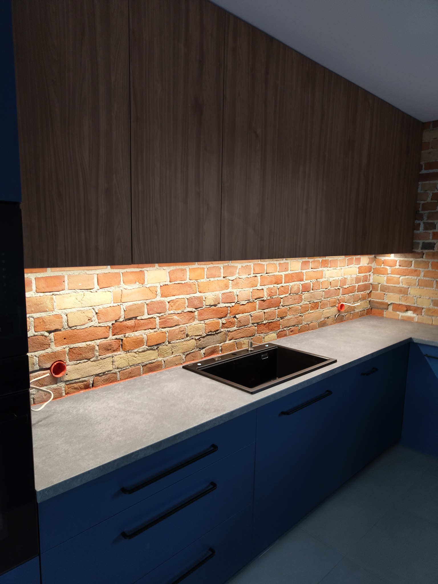 Nowoczesna kuchnia z ciemnoniebieskimi szafkami z długimi, czarnymi uchwytami, szarym blatem i ścianą wyłożoną cegłą podświetloną od góry, nad zlewem widoczne gniazdka elektryczne.