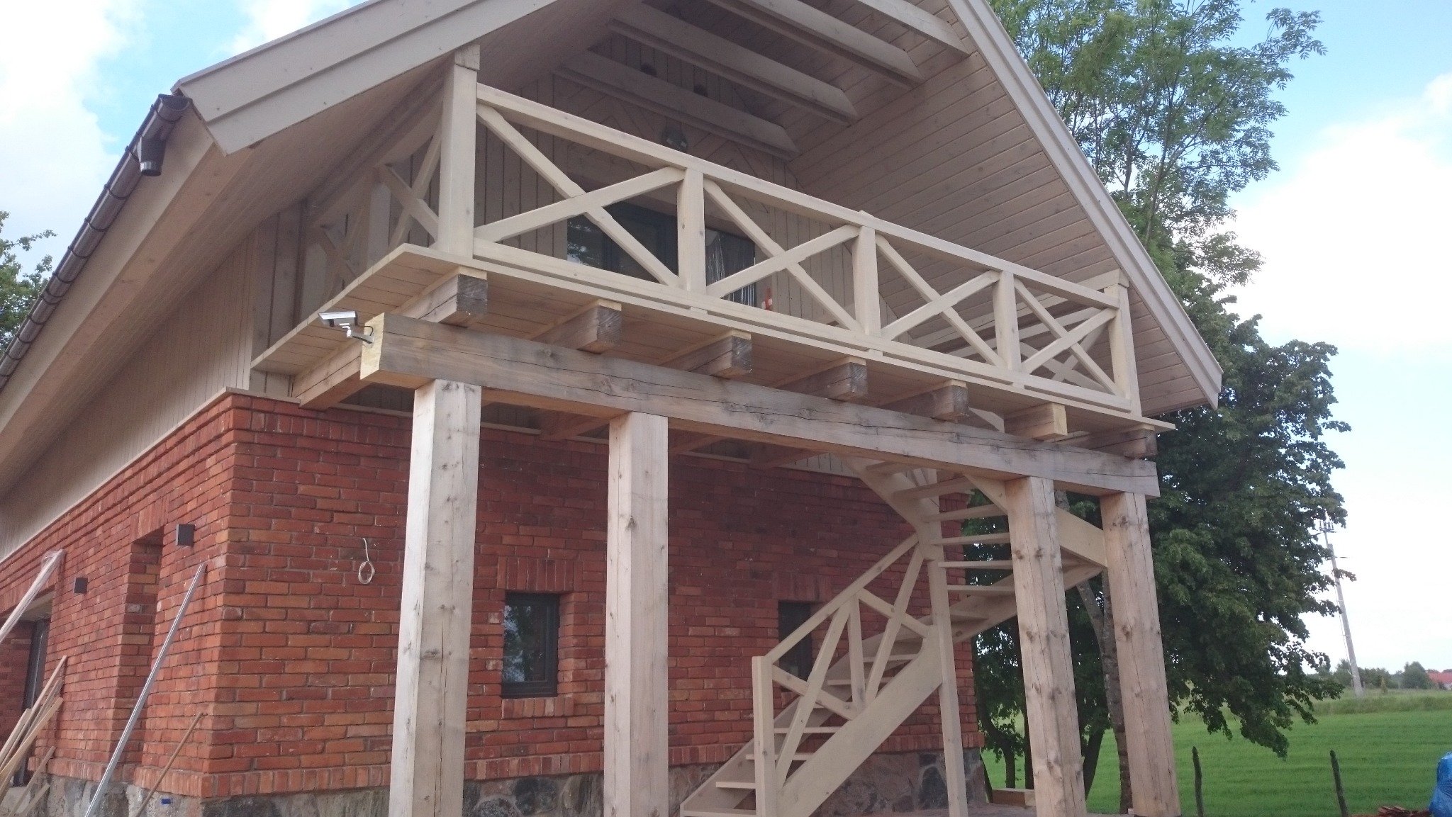 Dom z cegły z drewnianym tarasem na słupach i schodami, widok na zielony krajobraz w tle.