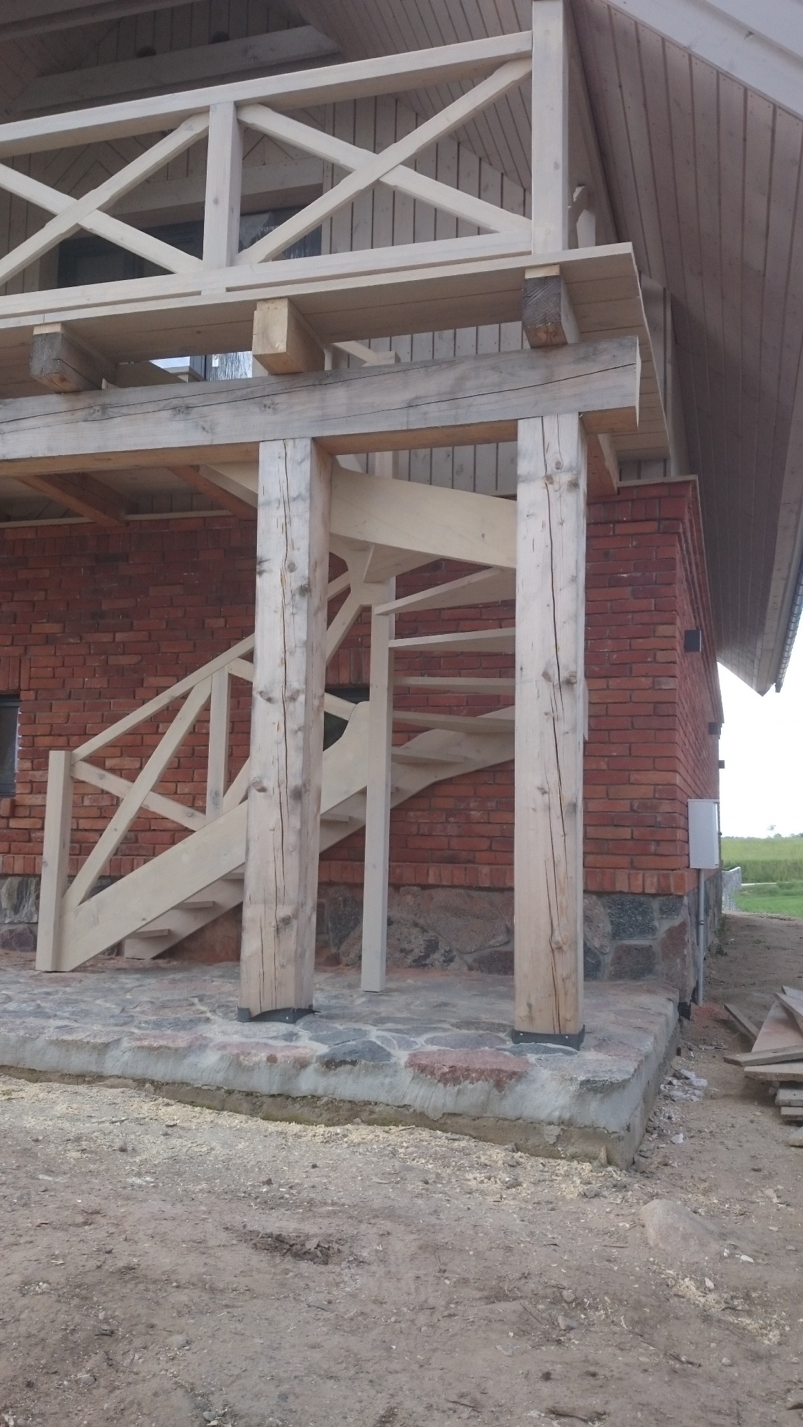 Surowe drewniane schody zewnętrzne z balustradą, prowadzące na drewniany taras domu z ceglaną elewacją, widoczne ślady obróbki drewna.