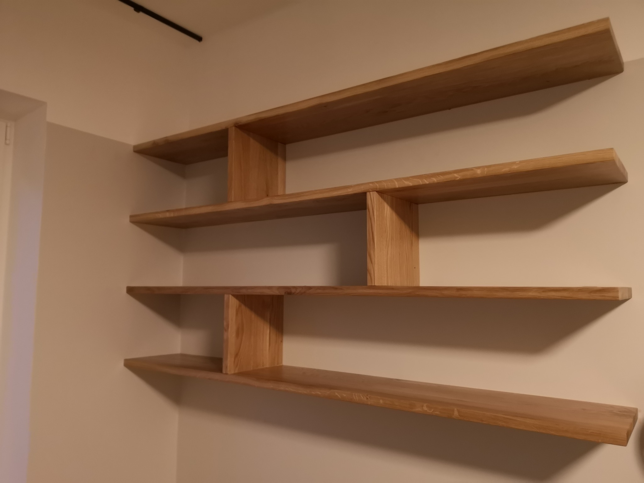 Asymetryczne, wiszące półki z jasnego drewna w minimalistycznym wnętrzu, mocowane do ściany, z widocznymi naturalnymi słojami drewna i subtelnym oświetleniem.