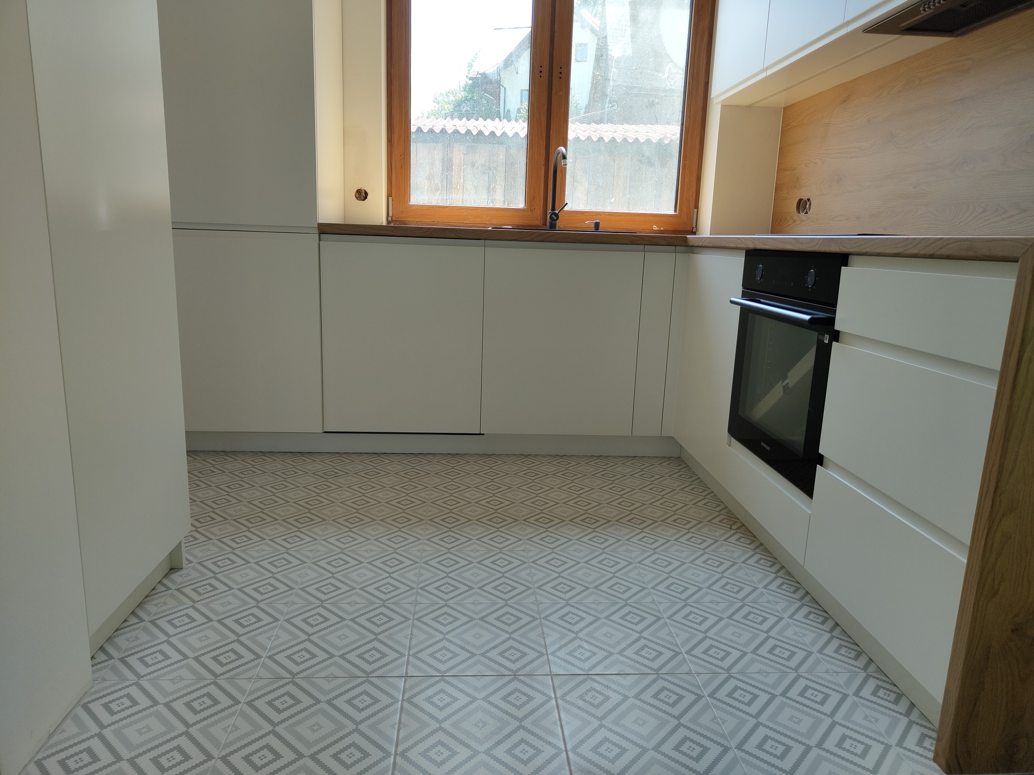 Jasna kuchnia z białymi frontami, drewnianym blatem i wzorzystą podłogą. Widoczny zlew pod oknem i piekarnik w zabudowie.