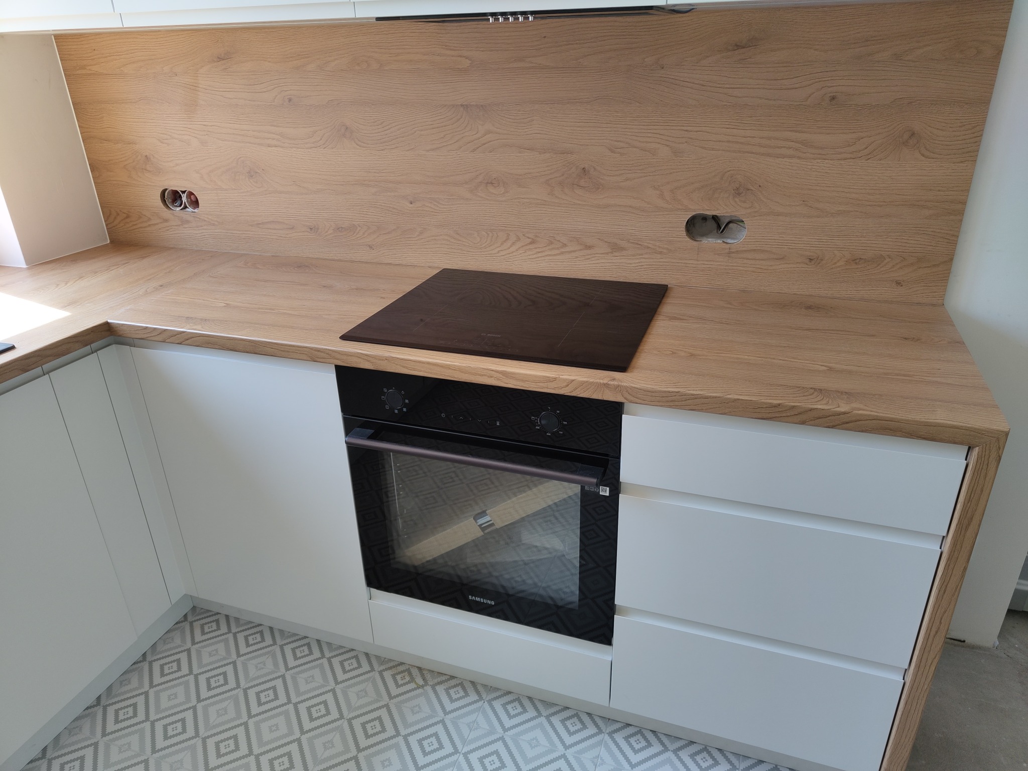 Nowoczesna kuchnia z białymi szafkami i blatem imitującym drewno, płyta indukcyjna wkomponowana w blat, widoczne gniazdka elektryczne nad blatem, wzorzysta podłoga.