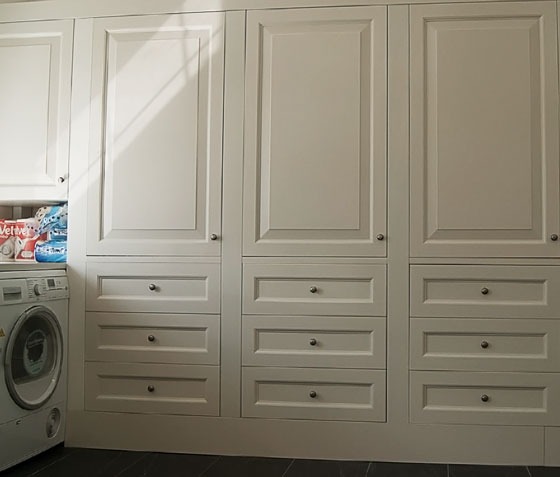 Biała zabudowa meblowa z szafkami i szufladami, wkomponowana w przestrzeń pralni obok pralki i zapasu środków czystości.